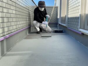 職人がバルコニーの床にローラーでグレーの防水塗料を丁寧に塗り広げている作業中の写真